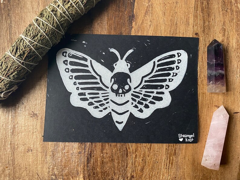May include: Black and white linocut print of a moth with a skull head. The print is on black paper and has the artist's signature "Strange Knot" in the bottom right corner.