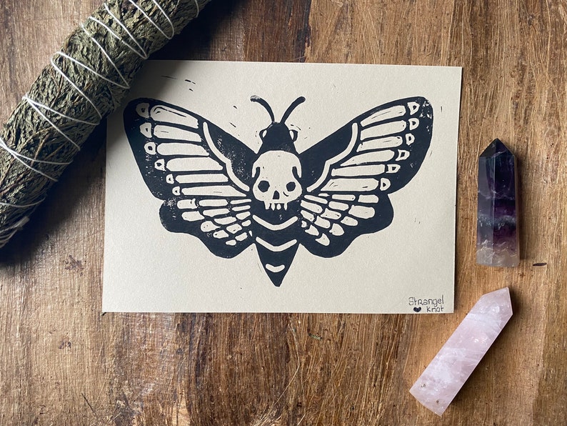 May include: Black and white linocut print of a moth with a skull head. The print is on a cream-colored paper and has the artist's signature "Branjel Kraft" in the bottom right corner.