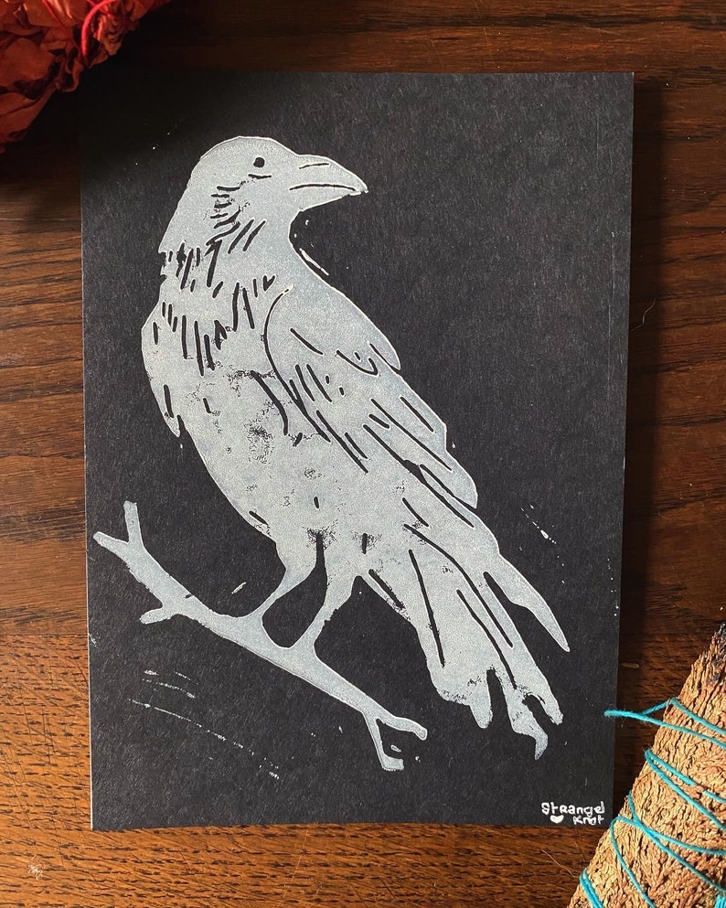 Peut inclure: Une silhouette blanche de corbeau perch&eacute; sur une branche sur un fond noir. L'image est une estampe linogravure avec la signature de l'artiste "Strange Knot" en bas &agrave; droite.