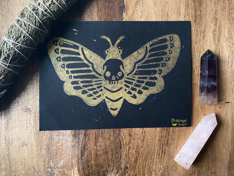 May include: A gold-colored print of a moth with a skull on its body, on a black background. The print is signed "Strange Knot" in the bottom right corner.