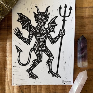 Peut inclure: Une impression linogravure en noir et blanc sur papier représentant un diable cornu et ailé tenant un trident. Le personnage a une queue et des marques de fourrure texturées. Le texte "Strange Knot" avec un petit cœur est imprimé en bas à droite.
