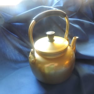 May include: A gold-colored metal tea kettle with a black handle and a black knob on the lid. The kettle is sitting on a blue fabric surface.