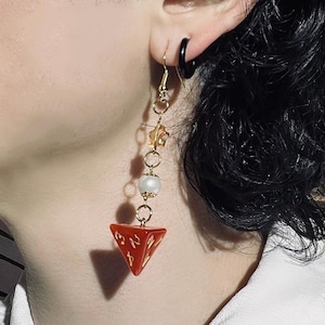 May include: A single gold-toned earring with a red, triangular dice charm. The earring features a pearl bead, clear crystal accents, and a gold-coloured ear hook. The dice charm has gold-coloured numbers.