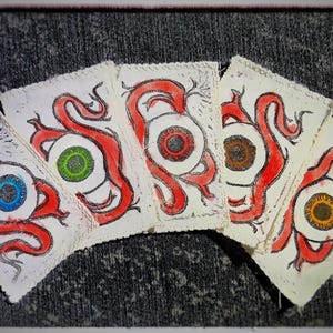 Könnte beinhalten: Fünf quadratische Karten mit handgezeichneten Illustrationen von Augen mit roten Adern. Jedes Auge hat eine andere Iris-Farbe: blau, grün, rot, braun und gelb. Die Karten sind fächerförmig angeordnet.