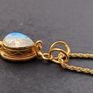 May include: A gold pendant necklace featuring a teardrop-shaped moonstone. The moonstone has a blue and white iridescent glow. The pendant is set in a gold bezel with intricate detailing, and hangs from a gold chain.