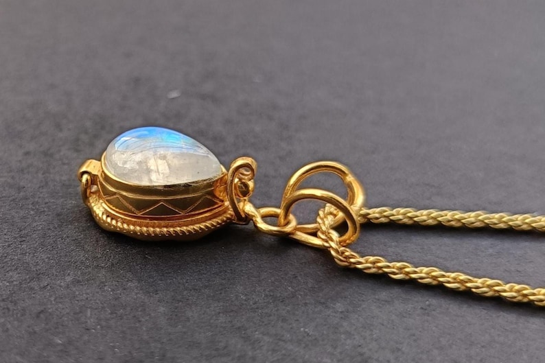 May include: A gold pendant necklace featuring a teardrop-shaped moonstone. The moonstone has a blue and white iridescent glow. The pendant is set in a gold bezel with intricate detailing, and hangs from a gold chain.