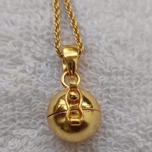Puede incluir: Un collar dorado con un colgante esférico y un cierre. El colgante cuelga de una fina cadena dorada. El collar se presenta sobre un fondo blanco texturizado.