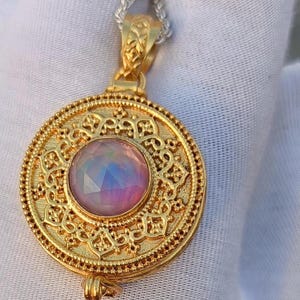 May include: A gold-colored pendant necklace with a round, ornate design. The pendant features a central, iridescent gemstone with pink, blue, and purple hues. The necklace has a delicate silver chain.