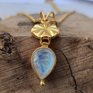 May include: A gold-colored pendant necklace featuring a teardrop-shaped moonstone. The moonstone has a blue and white iridescent glow. The pendant is attached to a delicate gold chain. The necklace is displayed on a wooden surface.
