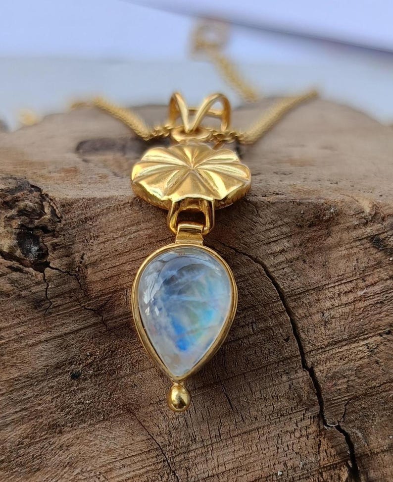 May include: A gold-colored pendant necklace featuring a teardrop-shaped moonstone. The moonstone has a blue and white iridescent glow. The pendant is attached to a delicate gold chain. The necklace is displayed on a wooden surface.