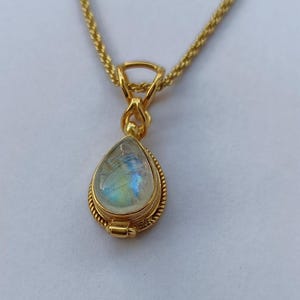 May include: A gold-colored necklace with a teardrop-shaped pendant. The pendant features a pearlescent, iridescent stone, possibly moonstone, set within a detailed, textured gold frame. The chain is a delicate, twisted rope design.