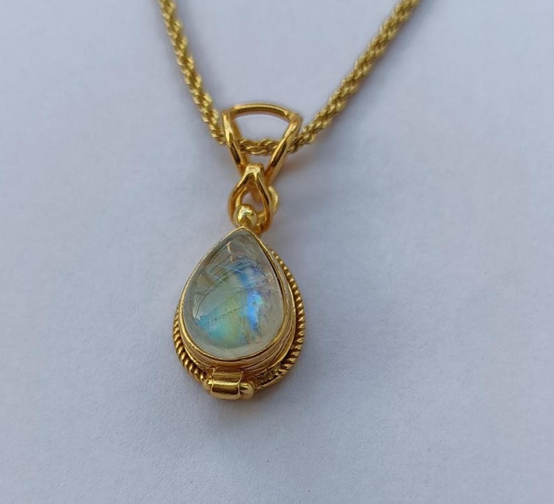 May include: A gold-colored necklace with a teardrop-shaped pendant. The pendant features a pearlescent, iridescent stone, possibly moonstone, set within a detailed, textured gold frame. The chain is a delicate, twisted rope design.