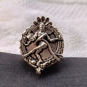 Puede incluir: Un colgante de plata con una representación detallada de una deidad hindú en una pose de baile, rodeado de intrincados elementos decorativos.