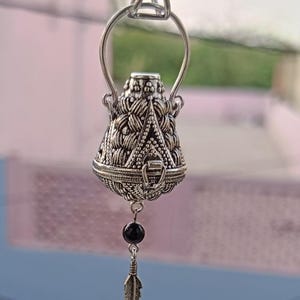 Op de afbeelding: Een zilverkleurige hangerketting met een gedetailleerde, patroonvormige lantaarnvormige bedel. De bedel heeft een kleine zwarte kraal en een veervormige hanger. De ketting hangt aan een delicate ketting, wat een elegante en bohemien uitstraling creëert.