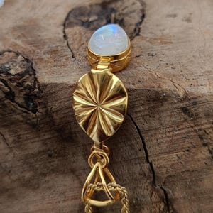 May include: A gold-colored pendant necklace featuring a pear-shaped, iridescent moonstone at the top. Below the moonstone is a gold, sunburst-patterned oval. The pendant hangs from a delicate gold chain, creating an elegant and eye-catching piece of jewelry.