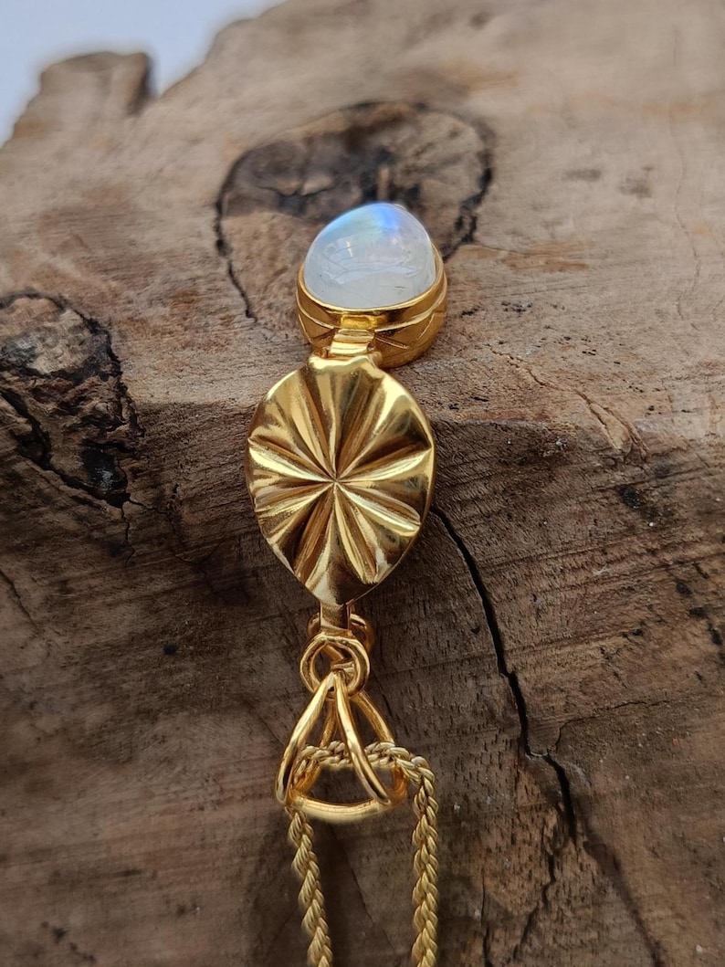 May include: A gold-colored pendant necklace featuring a pear-shaped, iridescent moonstone at the top. Below the moonstone is a gold, sunburst-patterned oval. The pendant hangs from a delicate gold chain, creating an elegant and eye-catching piece of jewelry.
