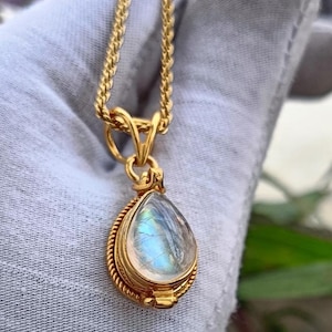 May include: A gold-colored necklace with a teardrop-shaped pendant. The pendant features a pearlescent, iridescent stone set within a detailed, rope-like border. The chain is a delicate, woven design.