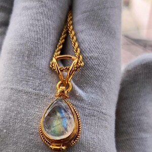 May include: A gold-colored necklace with a teardrop-shaped pendant. The pendant features a clear, iridescent stone with blue, green, and yellow hues. The necklace chain is a twisted rope design.