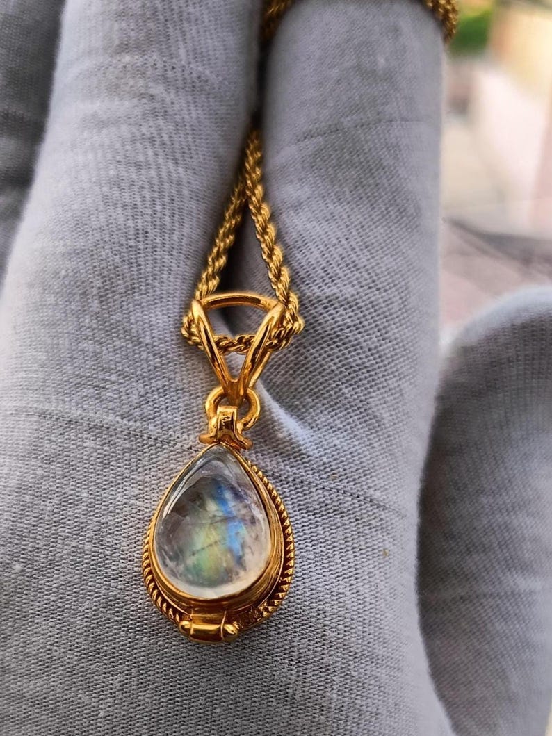 May include: A gold-colored necklace with a teardrop-shaped pendant. The pendant features a clear, iridescent stone with blue, green, and yellow hues. The necklace chain is a twisted rope design.
