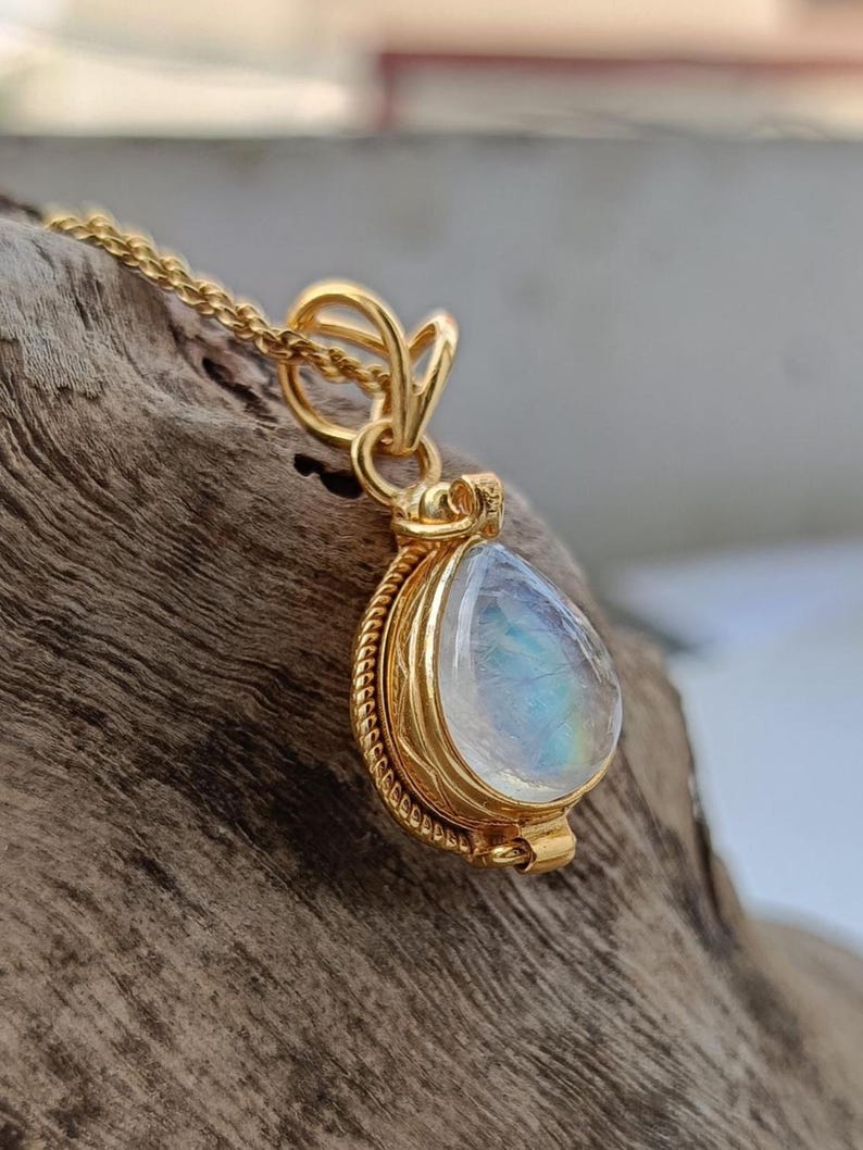 May include: A gold-colored necklace with a teardrop-shaped pendant. The pendant features a pearlescent, iridescent stone set in a detailed gold frame. The chain is delicate and the pendant is suspended from a decorative bail.