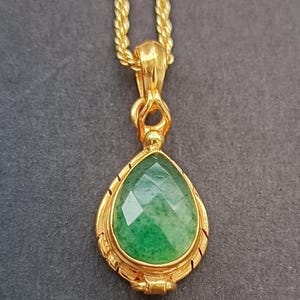 Op de afbeelding: Een goudkleurige hangerketting met een druppelvormige, gefacetteerde groene edelsteen. De edelsteen is gezet in een goudkleurige bezel met een decoratieve rand. De ketting is een goudkleurige touwstijl.