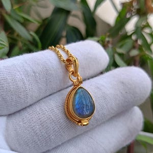 Puede incluir: Collar con colgante dorado con una piedra de labradorita en forma de lágrima, con tonos azules e iridiscentes. La piedra está engastada en un marco dorado texturizado y detallado, suspendida de una fina cadena. La joya se muestra sobre un fondo blanco.