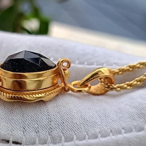 May include: Gold-toned pendant necklace featuring a faceted, dark blue gemstone. The pendant is encased in a gold setting with intricate detailing, suspended from a twisted gold chain. The necklace is displayed on a white surface.