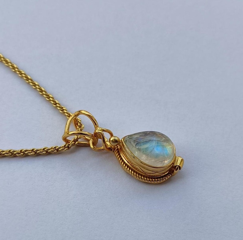 May include: A gold-colored necklace with a teardrop-shaped pendant. The pendant features a pearlescent, iridescent stone set in a gold bezel. The chain is a delicate, woven design. The necklace is displayed on a light background.