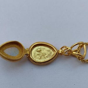May include: A gold-colored pendant necklace with a teardrop-shaped locket. The locket is open, revealing a pearlescent stone and a detailed interior. The necklace chain is a delicate gold color.