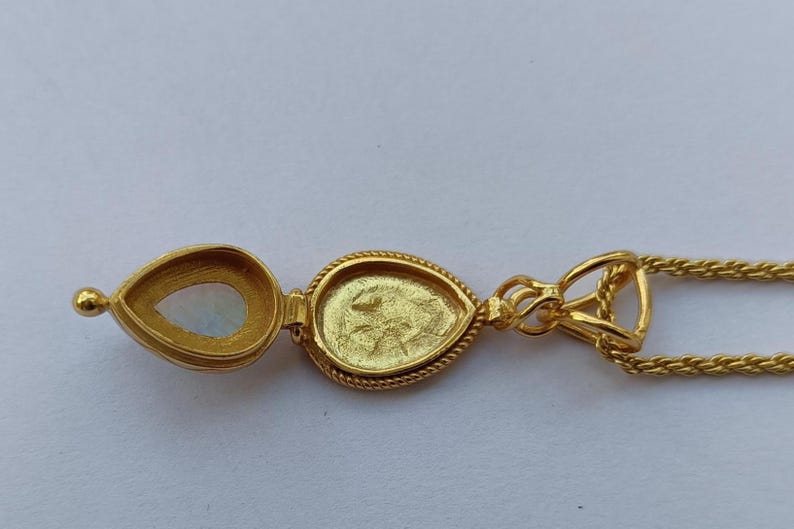 May include: A gold-colored pendant necklace with a teardrop-shaped locket. The locket is open, revealing a pearlescent stone and a detailed interior. The necklace chain is a delicate gold color.