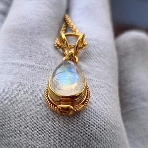 May include: A gold-colored pendant necklace featuring a pear-shaped, iridescent moonstone. The moonstone is set in a detailed gold bezel with a rope-like border. The pendant hangs from a delicate gold chain.
