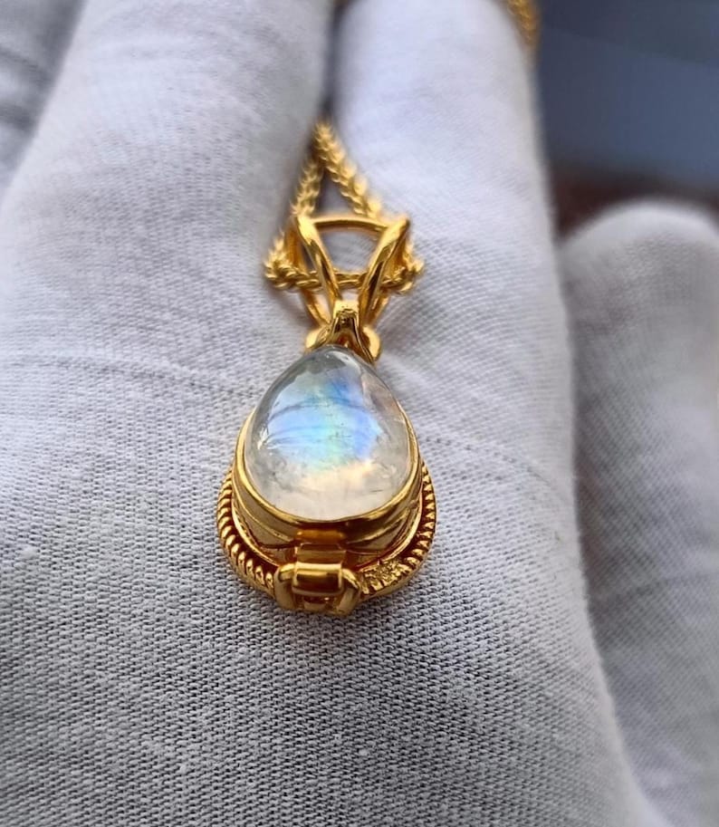 May include: A gold-colored pendant necklace featuring a pear-shaped, iridescent moonstone. The moonstone is set in a detailed gold bezel with a rope-like border. The pendant hangs from a delicate gold chain.