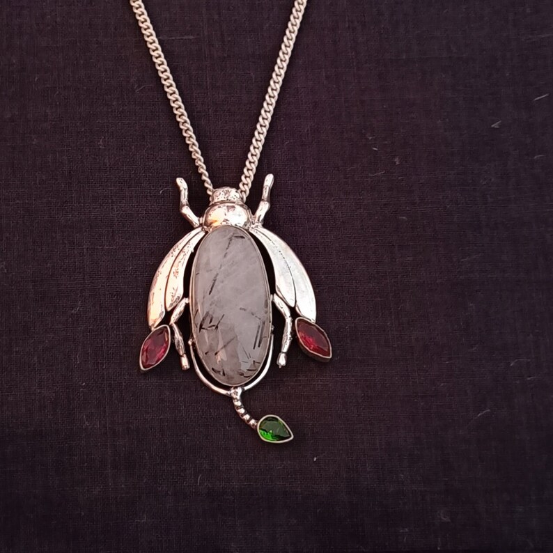 May include: A silver necklace with a pendant shaped like a beetle. The beetle has a clear quartz stone in the center and red and green gemstones on the sides.