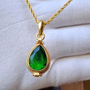 May include: A gold-colored necklace with a teardrop-shaped pendant. The pendant features a faceted, emerald-green gemstone set within a detailed gold frame. The chain is a twisted rope design.