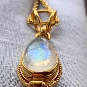 May include: A gold-colored pendant with a teardrop-shaped, iridescent gemstone. The pendant has a detailed, ornate design with a textured border and a decorative bail. The gemstone displays a play of colors, including blue and yellow.