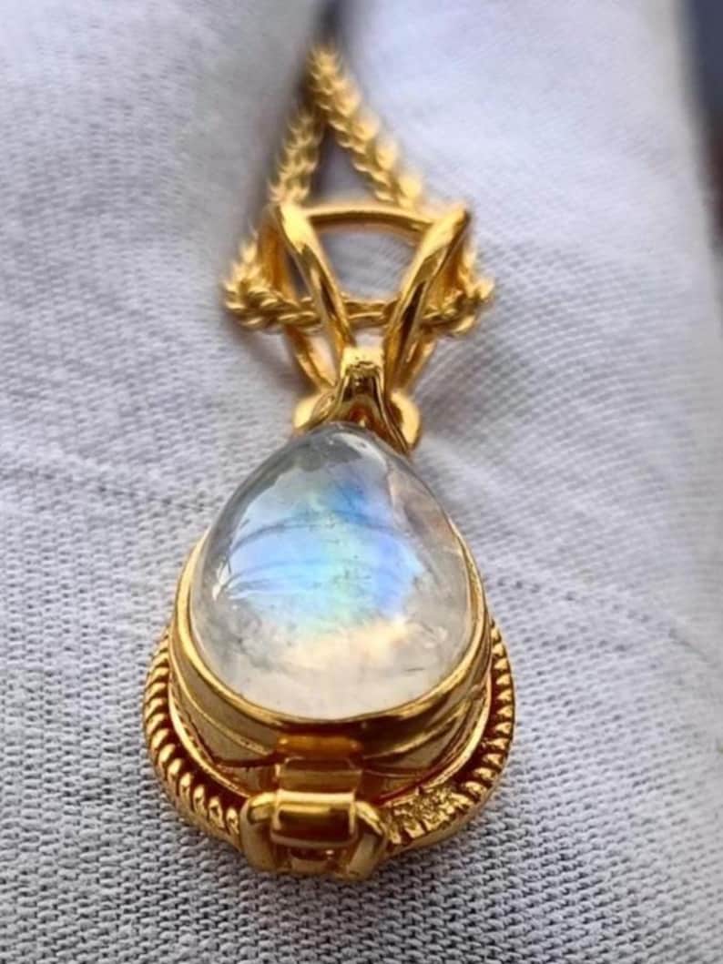 May include: A gold-colored pendant with a teardrop-shaped, iridescent gemstone. The pendant has a detailed, ornate design with a textured border and a decorative bail. The gemstone displays a play of colors, including blue and yellow.