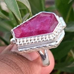 Puede incluir: Un anillo de plata con una piedra de rubí rosa engastada en un diseño en forma de ataúd. El anillo está decorado con un borde de cuentas y una banda texturizada.
