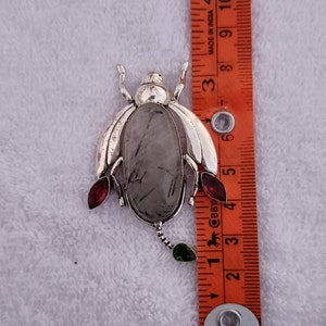 May include: Silver beetle brooch with a clear quartz stone and red and green accents. The brooch is approximately 2 inches long.