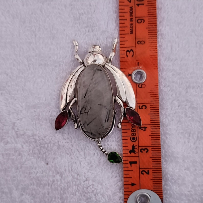 May include: Silver beetle brooch with a clear quartz stone and red and green accents. The brooch is approximately 2 inches long.