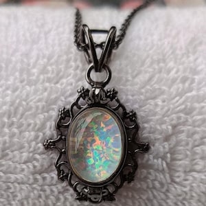 May include: A black metal pendant necklace with an oval-shaped iridescent stone. The stone is set within a decorative, ornate frame. The necklace is displayed on a white, textured surface.