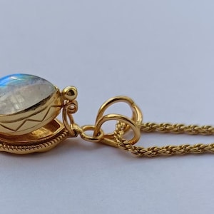 May include: A gold-colored pendant necklace featuring a round, iridescent gemstone. The gemstone is set in a detailed, decorative gold setting. The necklace chain is a delicate, twisted rope design.