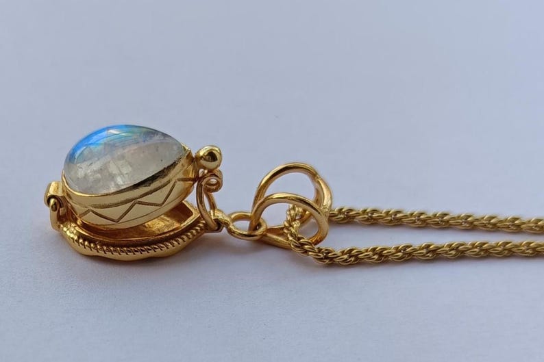 May include: A gold-colored pendant necklace featuring a round, iridescent gemstone. The gemstone is set in a detailed, decorative gold setting. The necklace chain is a delicate, twisted rope design.