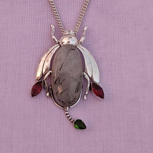May include: A silver pendant necklace featuring a grey gemstone with a silver bug design. The bug has red and green accents.