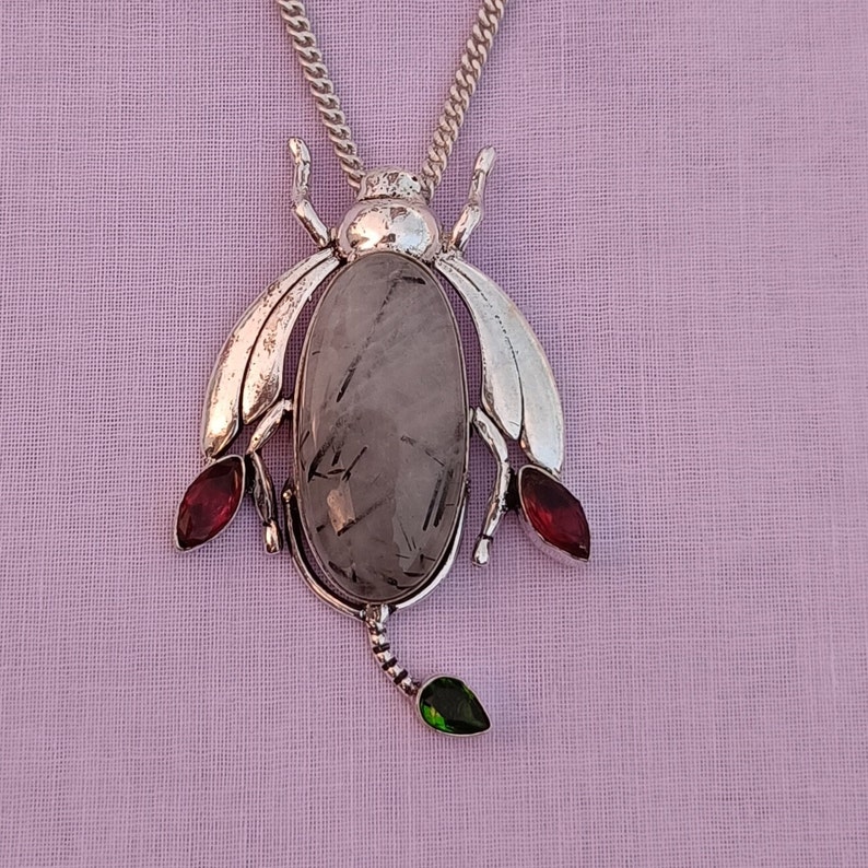 May include: A silver pendant necklace featuring a grey gemstone with a silver bug design. The bug has red and green accents.