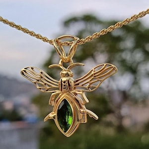 May include: A gold-colored bee pendant with a green gemstone center, suspended from a matching chain. The bee's wings are intricately detailed, and the pendant is set against a blurred outdoor background.