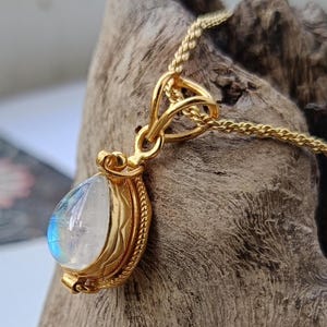 May include: A gold-colored pendant necklace featuring a pear-shaped, iridescent moonstone. The moonstone is set in a detailed, ornate gold bezel. The chain is a delicate, woven design.