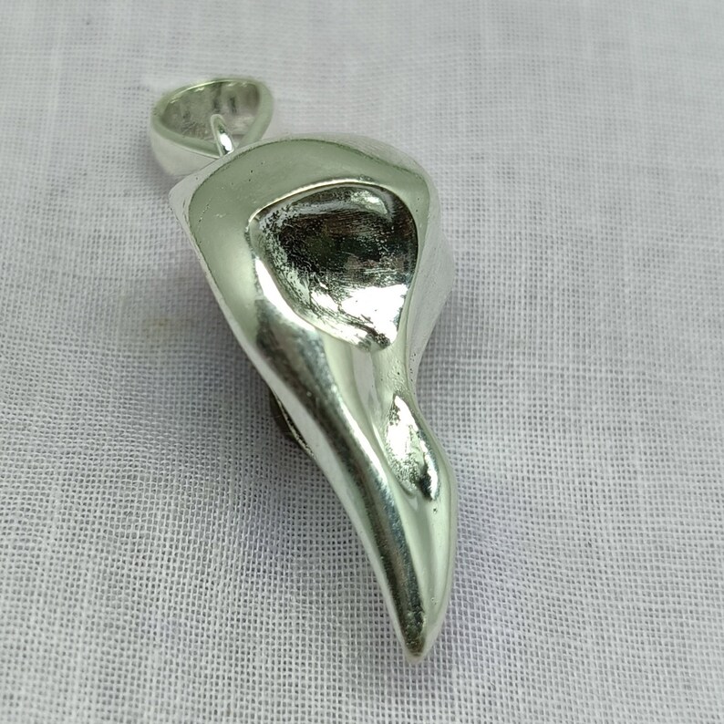 Puede incluir: Un colgante de plata con forma de cabeza de p&aacute;jaro con un ojo hueco. El colgante es liso y brillante.