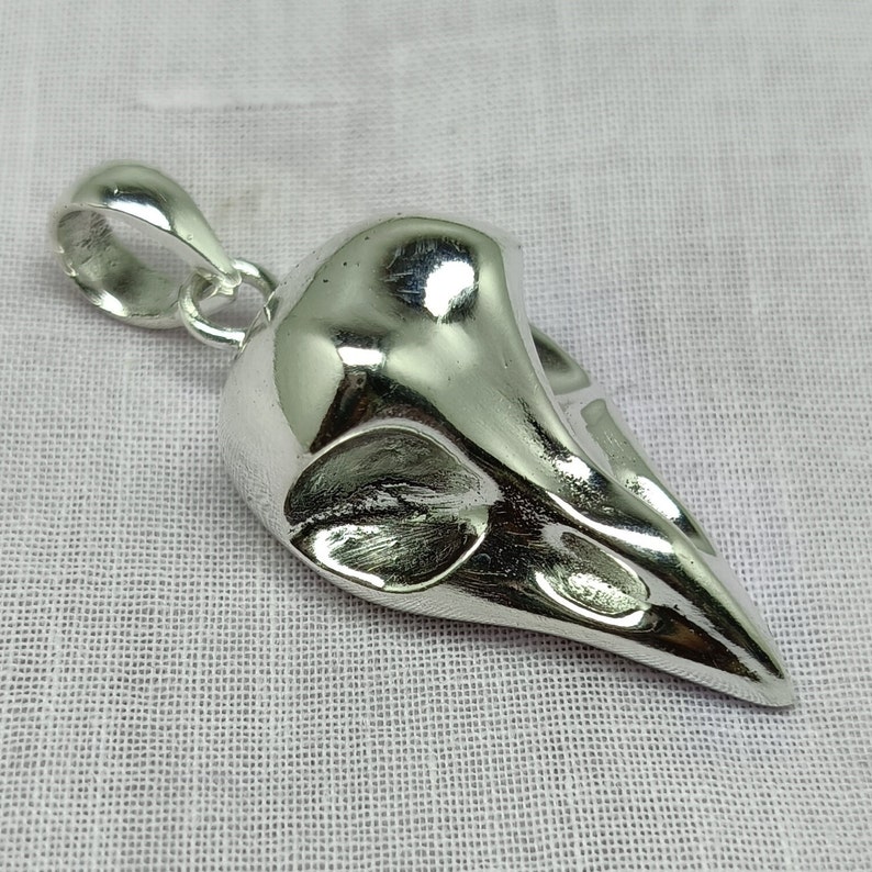 Puede incluir: Un colgante de plata con forma de cr&aacute;neo de p&aacute;jaro. El colgante tiene un bucle en la parte superior para sujetarlo a una cadena.