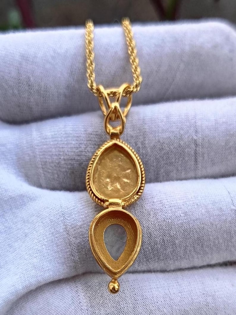 May include: A gold-colored locket necklace with a teardrop-shaped pendant. The pendant opens to reveal a small compartment. The necklace chain is a twisted rope design. The locket has a decorative rope-like border.