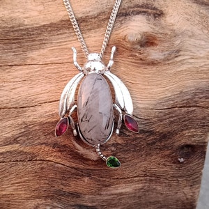 May include: A silver pendant necklace featuring a beetle design with a clear quartz stone and red and green accents.
